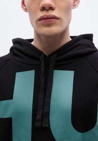 Μαύρο φούτερ με κουκούλα που διαθέτει ένα εντυπωσιακό γραφικό σε τιρκουάζ χρώμα. Έχει ρυθμιζόμενο σκοινάκι στη λαιμόκοψη και απαλό, υφασμάτινο υλικό με υφή. Κοντινή όψη.