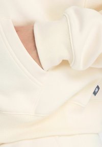 Sweatshirt de couleur crème avec des poignets côtelés et une poche kangourou, en tissu lisse et avec des détails de couture subtils sur les coutures.