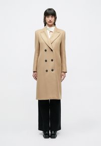 Μπεζ παλτό με διπλή σειρά κουμπιών κατασκευασμένο από μαλλί, με μαύρα κουμπιά, γιακάδες με εγκοπές και πλαϊνές τσέπες· συνδυασμένο με μαύρο παντελόνι με φαρδιά μπατζάκια και μπότες.