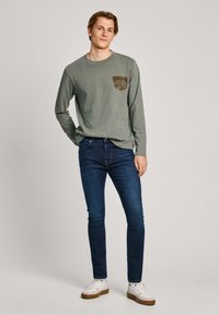 Dolga rukava olivno zelena majica z žepom na prsih, kombinirana s slim-fit temno modrimi kavbojkami in belimi supergami z rjavimi detajli.