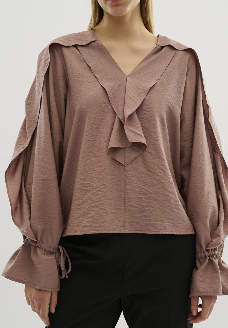 Blusa color rosa con scollo a V, dettagli a strati con volant e maniche oversize. Realizzata in tessuto testurizzato con polsini arricciati e nastri.