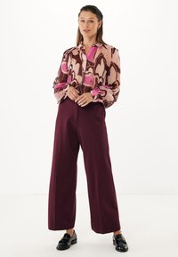 Kvinna som står klädd i en genomskinlig blommig blus i rosa och brunt, högmidjade vida maroonbyxor och svarta loafers på vit bakgrund.