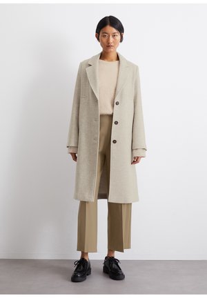 Cappotto di lana color beige chiaro con rever a cappotto, design monopetto, tre bottoni e tasche laterali. Indossato sopra un maglione di lana beige e pantaloni beige.