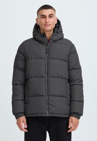 Grå pufferjakke med hætte, med et quiltet design, frontlynlås og elastiske manchetter, båret af en person foran en neutral baggrund.