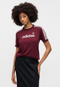 Bordeauxrode korte mouw T-shirt met wit "adidas"-logo en drie witte horizontale strepen op de schouders, gecombineerd met een rok met zwarte elastische tailleband.