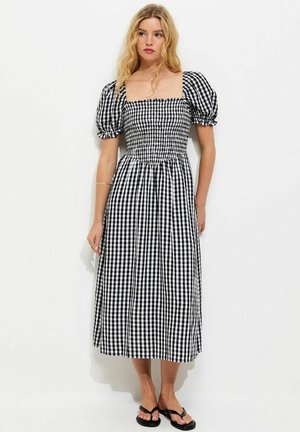 Kobieta w czarno-białej sukience midi w kratę gingham z bufkami, kwadratowym dekoltem i czarnych japonkach, stojąca na białym tle.