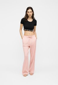 Γυναίκα στέκεται ξυπόλυτη φορώντας μαύρο κοντό μπλουζάκι με λωρίδα λογότυπου και φαρδιά ροζ παντελόνια με κορδόνι σε λευκό φόντο.