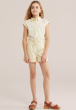 Ung pige står iført en sart gul, ærmeløs skjorte med knapper og matchende shorts med små frugtprint samt brune sandaler.