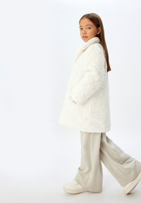 Cappotto bianco, texturizzato, simile a pelliccia, con colletto e tasche laterali, indossato sopra pantaloni beige chiaro a gamba larga e sneakers bianche.