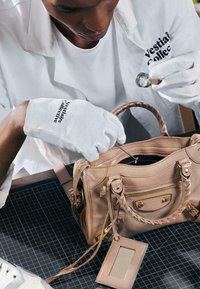 Personne portant des gants blancs examine un sac à main en cuir beige avec une loupe sur un tapis à grille noire.