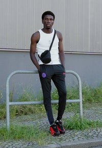 Hvid ærmeløs tanktop, sorte Nike træningsbukser med røde og blå detaljer, sort crossbody taske og røde sneakers. Står ved et metal rækværk.