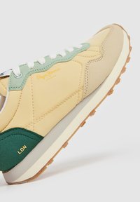 Basket de Pepe Jeans avec un design jaune, vert et beige, dotée d'un dessus texturé, de lacets blancs et d'une semelle en caoutchouc. Détail du logo doré présent.