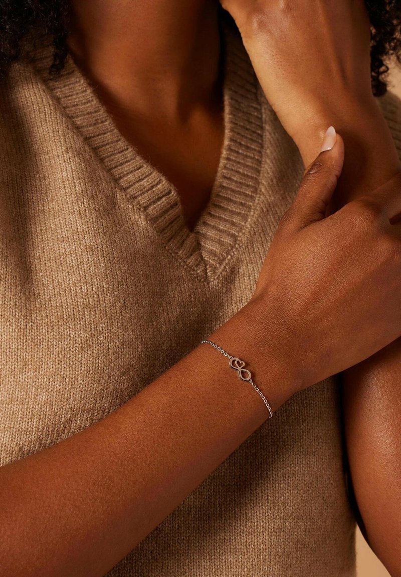 Zilveren schakelarmband met een infinity-symbool als sluiting, gedragen op een middengouden huidtint, tegen een gestructureerde beige gebreide trui.