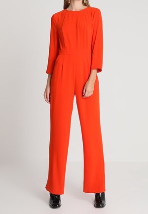 Vrouw draagt een feloranje jumpsuit met lange mouwen, wijde pijpen en zwarte hakken tegen een effen achtergrond.