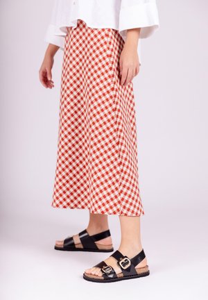 Persona que lleva pantalones anchos a cuadros rojos y blancos, camiseta blanca de manga larga y sandalias negras con hebillas doradas, de pie sobre un suelo blanco.