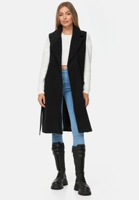 Schwarze lange Weste aus dickem Stoff mit Spitzkragen. Tragen über einem weißen kurzen Pullover und kombiniert mit blauen Skinny-Jeans und schwarzen Combat-Stiefeln.