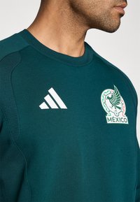 Τιρκουάζ αθλητική μπλούζα με στρογγυλή λαιμόκοψη, που φέρει το λευκό λογότυπο της Adidas στο αριστερό μέρος του στήθους και το πράσινο και λευκό έμβλημα της εθνικής ομάδας του Μεξικού.