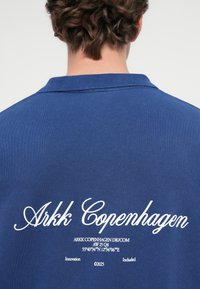 Sötétkék póló, amely puha tapintású, a hátsó részén fehér "Arkk Copenhagen" logóval és részletes grafikával, beleértve a koordinátákat.