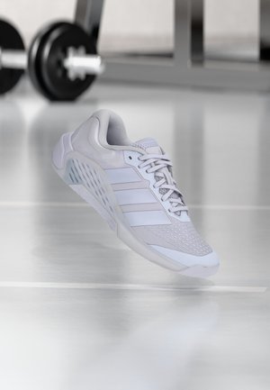 Zapatilla deportiva blanca con malla y superposiciones sintéticas, cordones atados, flotando sobre un suelo reflectante con equipo de gimnasio desenfocado en el fondo.