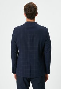 Marinblå blazer med rutmönster, med en enda slits, spetsiga reverser och knappdetaljer på ärmarna. Slen tygstruktur.