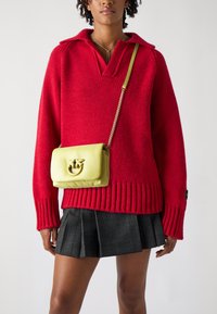 Maglione rosso a maglia con colletto e orlo a costine, abbinato a una gonna plissettata a quadri nera. Una borsa a tracolla in pelle gialla presenta un logo in tono oro.