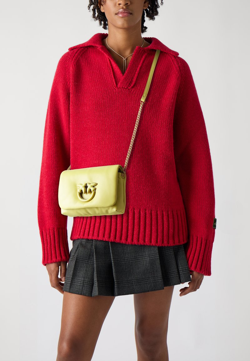 Maglione rosso a maglia con colletto e orlo a costine, abbinato a una gonna plissettata a quadri nera. Una borsa a tracolla in pelle gialla presenta un logo in tono oro.