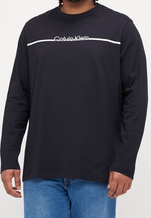 Mężczyzna ubrany w czarną koszulę Calvin Klein z długim rękawem, z białym paskiem nad logo, oraz niebieskie dżinsy, stojący na jednolitym tle.