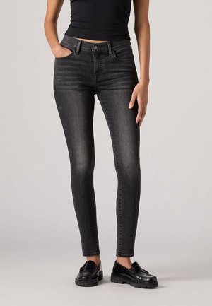Femme portant un jean skinny noir délavé, un débardeur noir et des mocassins noirs, debout avec une main dans la poche.