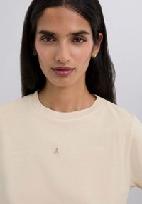 Camiseta de algodón beige con cuello redondo y un pequeño emblema de calavera y huesos cruzados en el pecho. Textura suave y ajuste casual.