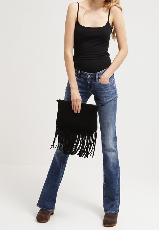 Femme portant un débardeur noir et un jean bleu, tenant une pochette noire à franges, debout avec une main dans la poche.