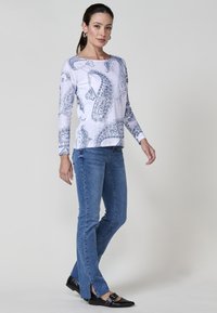 Mujer de pie vestida con una blusa blanca de manga larga con estampados de paisley azul, jeans azules con aberturas frontales y zapatos planos negros de punta.