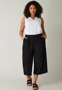 Zwarte cropped broeken met een gladde textuur, voorzien van een brede elastische tailleband, en een witte mouwloze top met V-hals. Draagt zwarte sandalen.