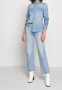 Chemise en denim bleu clair avec un devant boutonné, deux poches poitrine et des manches longues, associée à un jean large bleu clair et des bottines blanches.