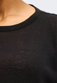 Κοντινό πλάνο ενός ατόμου που φοράει μαύρο μπλουζάκι με στρογγυλή λαιμόκοψη σε απλό φωτεινό φόντο.