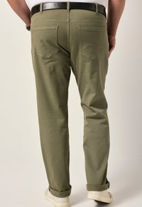 Pantalones verde oliva con pierna recta, que presentan una textura sutil, dos bolsillos traseros con detalles de costura y puños enrollados sobre zapatillas blancas.