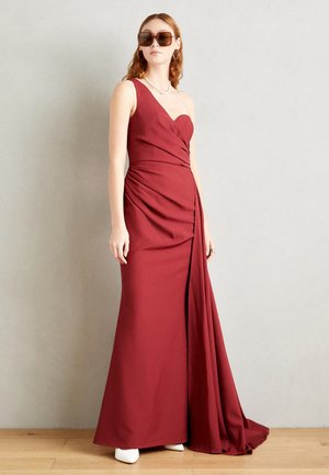 Robe rouge à une épaule avec des détails drapés, coupe longue jusqu'au sol, décolleté asymétrique et tissu lisse, assortie à des chaussures blanches et des lunettes de soleil.