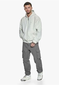 Lys grå zip-up hoodie, cargo bukser i mørkegrå og hvide sneakers. Hoodie har en afslappet pasform og frontlommer. Casual streetwear stil.