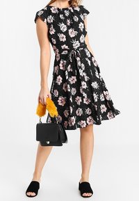 Sukienka w kwiatowy wzór z krótkim rękawem, czarne tło, różowe kwiaty, taliowany fason i rozkloszowany dół. Uzupełniona o czarną torebkę z żółtym pomponem.