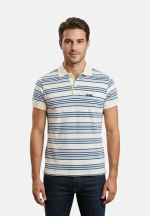 Uomo che indossa una polo a righe bianche e azzurro chiaro con colletto e jeans scuri, in piedi davanti a uno sfondo bianco uniforme.