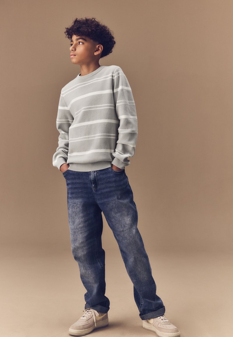 Felpa grigia a righe con maniche lunghe, abbinata a jeans blu larghi e sneakers di colore chiaro. Outfit semplice e casual.