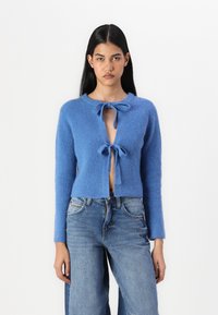 Cardigan bleu en tricot à manches longues, à ouverture sur le devant, avec deux liens au niveau du cou. Porté par-dessus un jean bleu taille haute. Tissu texturé.