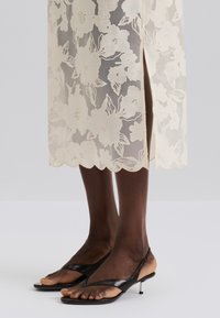 Transparenter, cremefarbener Spitzenrock mit Blumenmuster, gewelltem Saum und seitlichem Schlitz. Kombiniert mit schwarzen Leder-Sandalen mit Zehensteg und kleinem Absatz.