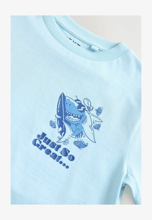 Jasnoniebieska bawełniana koszulka z grafiką rekina w ciemniejszym niebieskim, okularami przeciwsłonecznymi oraz napisem "Po prostu wspaniale..." z ilustracjami małych rybek.