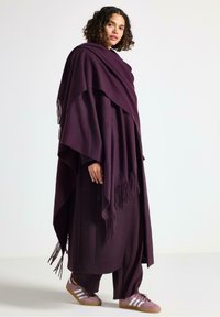Poncho viola con bordi frangiati appoggiato su un vestito coordinato, tessuto texturizzato, abbinato a sneakers rosa chiaro con strisce bianche.
