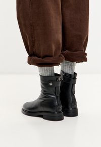 Botas de cuero negras hasta el tobillo con cremalleras laterales y correas, combinadas con pantalones marrones arremangados y calcetines de canalé grises sobre un fondo blanco.