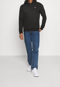 Svart hoodie med känguruficka och logotyp, tillsammans med blå jeans och vita sneakers. Slät tyg, avslappnad passform och enkel design.