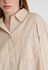 Close-up van een persoon in een beige geruite shirt met knopen, een borstzak, een ringoorbel en zichtbare haren op de huid.