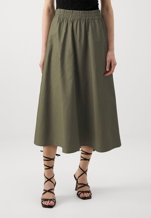 Kvinde iført olivengrøn midi-nederdel med elastisk taljebånd og sorte højhælede sandaler med remme, stående mod en ensfarvet baggrund.