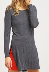Tunic a maniche lunghe grigia con scollatura rotonda e spaccature laterali, realizzata in un tessuto leggero e elastico. Indossata sopra una gonna rossa.