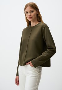 Felpa verde oliva con scollo rotondo, maniche lunghe e finitura testurizzata. Abbinata a jeans bianchi, con le mani infilate nelle tasche.